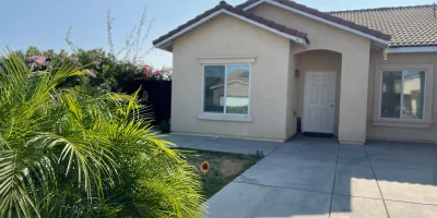 Exterior of a beige single-story care home with tiled roof, white door, large windows, and lush green plants in Fresno, California.